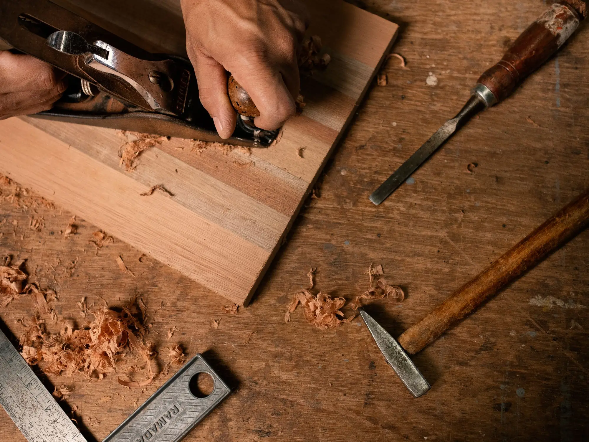 Handen bewerken een plank met een schaaf, met gereedschap ernaast.